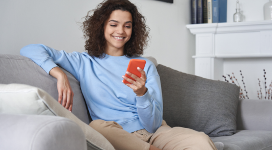 Mujer joven y sonriente revisando su teléfono celular mientras descansa cómodamente en el sofá de su sala.