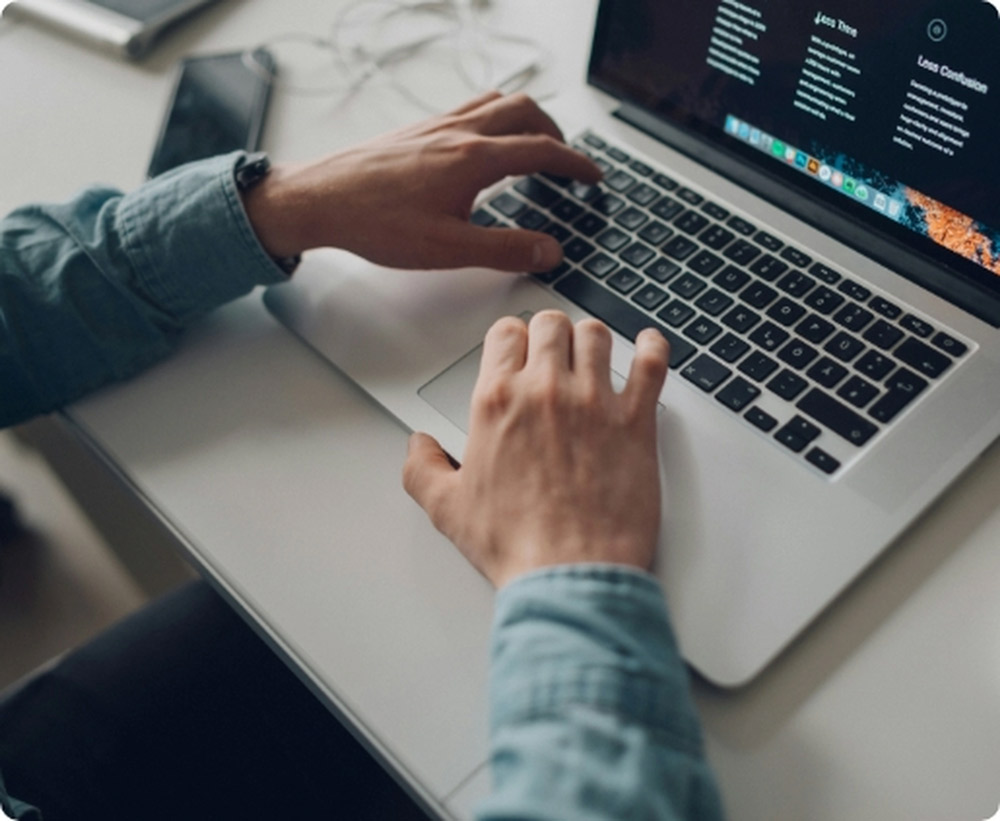Vista cenital de manos masculinas trabajando en el teclado de una laptop plateada; la pantalla muestra código de programación y texto en un entorno de oficina minimalista.