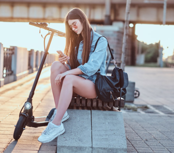 Mujer joven revisando su smartphone en la calle junto a un scooter eléctrico durante el atardecer.