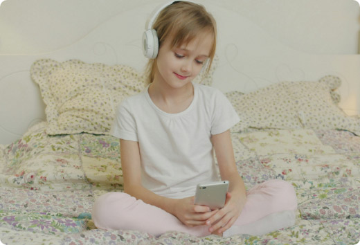 Niña sentada en su cama usando audífonos inalámbricos mientras consume contenido multimedia en un smartphone.