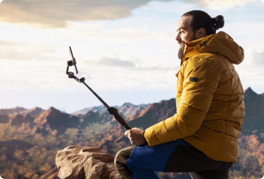 Hombre usando un smartphone con palo selfie en la montaña para grabar video o tomar fotos durante una actividad al aire libre.