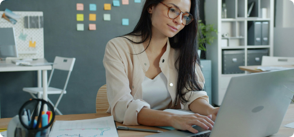 Mujer con gafas trabajando concentrada frente a su notebook en una oficina moderna y luminosa, con notas adhesivas y organizadores en el fondo.