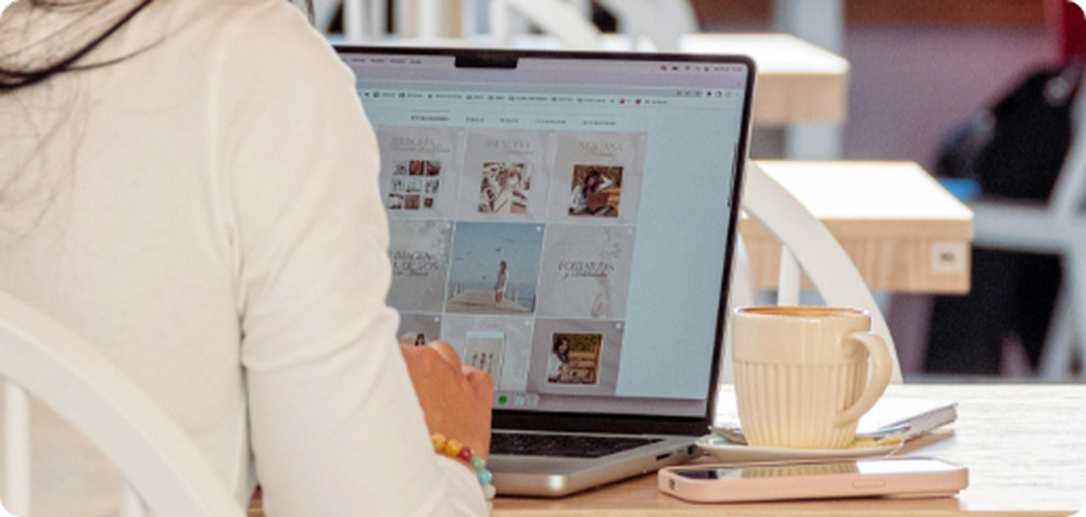 Vista sobre el hombro de una persona navegando por una galería de imágenes en un notebook de uso cotidiano en una cafetería, con una taza de café y un celular sobre la mesa.